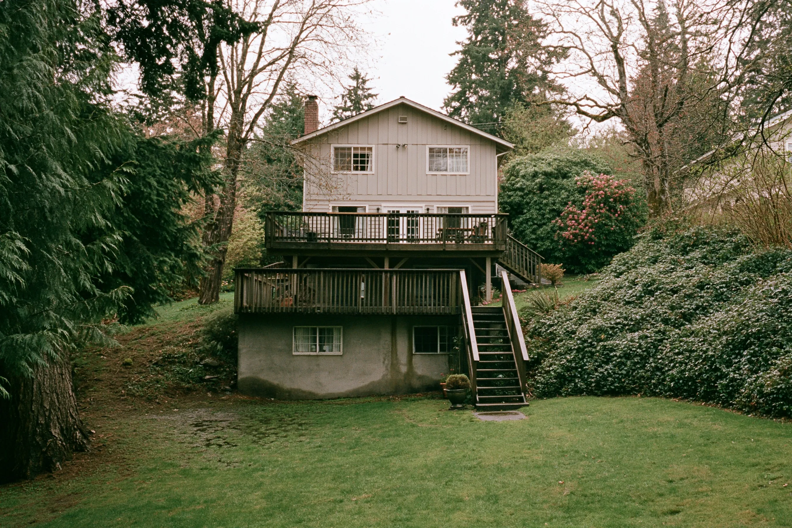 Rear of a two-story house on a sloped lot with a daylight basement, a concrete lower wall, a deck above and tall evergreens behind